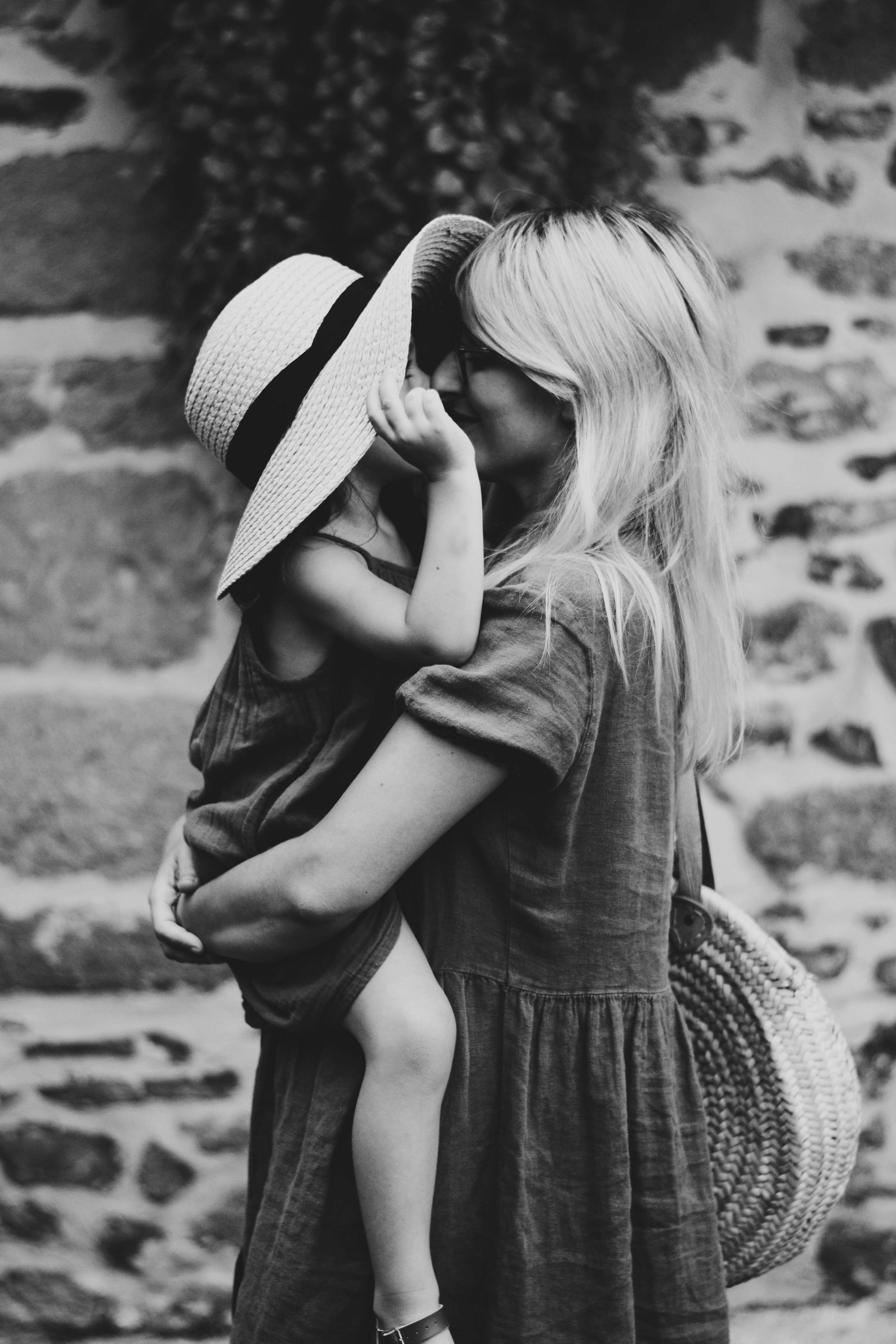 A black and white photograph of a woman with long blonde hair holding a young child wearing a wide-brimmed straw hat, both in casual clothing, with a woven basket visible and a blurred stone wall background.