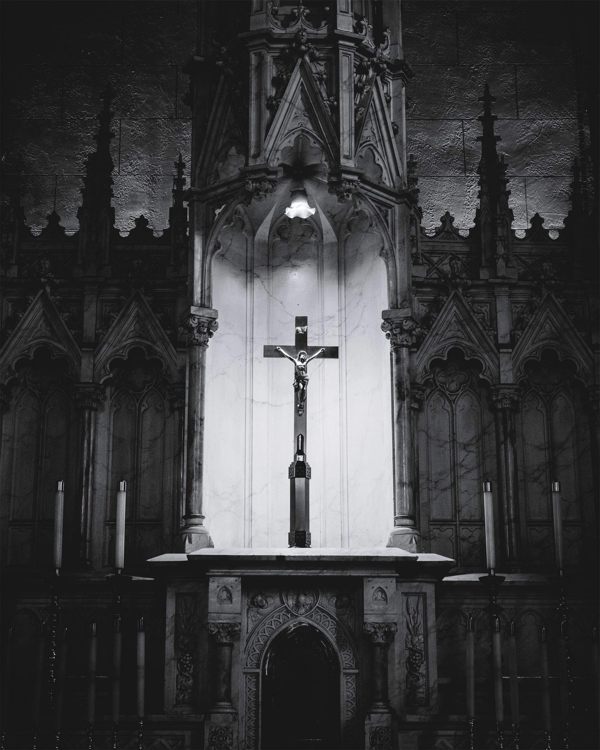 A black and white photograph of an ornate Gothic church altar featuring elaborate stone carved arches, decorative columns with detailed capitals, a crucifix mounted on the wall above a marble altar table, and candles positioned on either side, all illuminated by dramatic lighting that emphasizes the architectural details.
