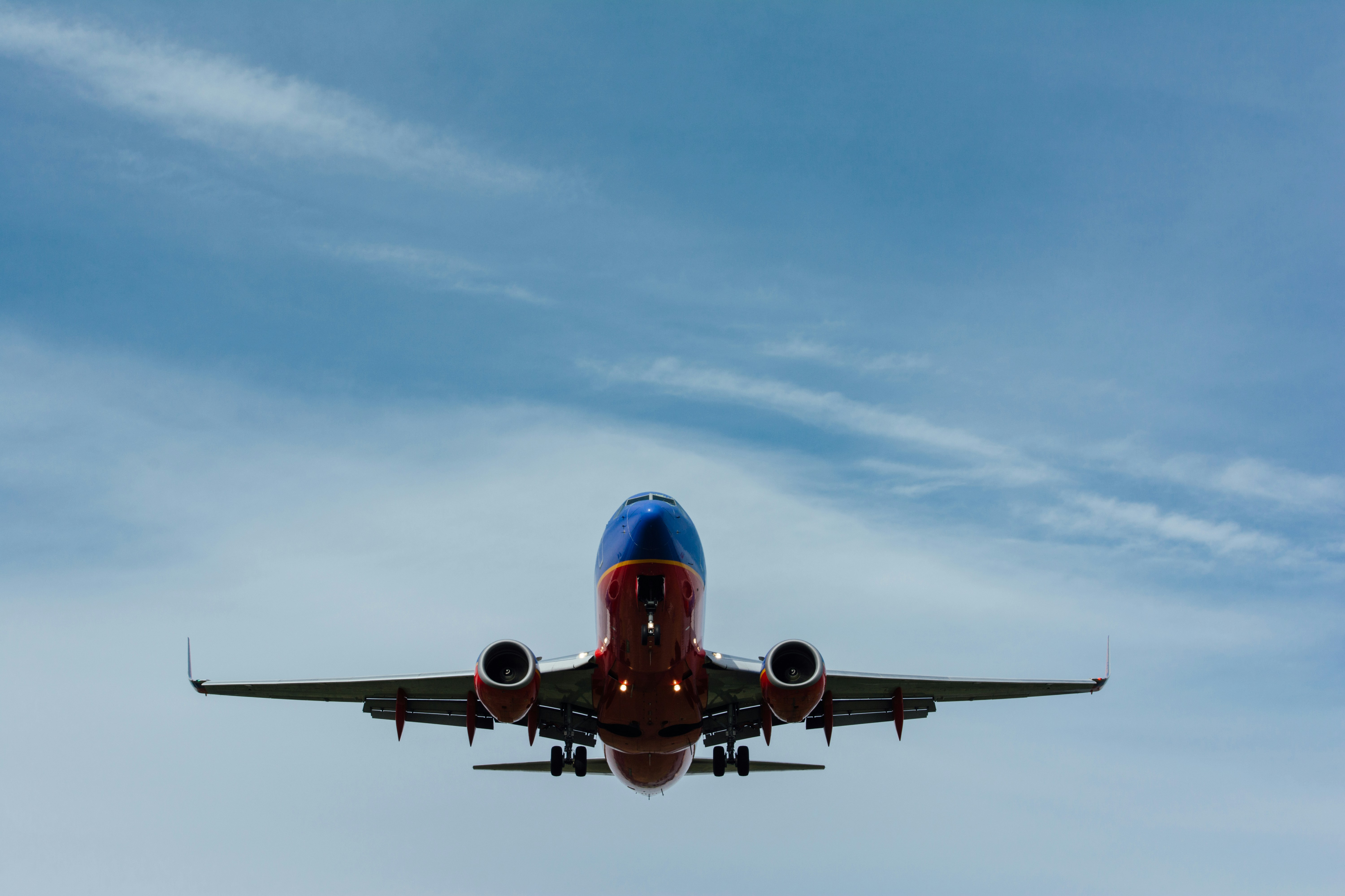 A commercial airplane flying through a partly cloudy blue sky, photographed from below showing the aircraft's underside with its extended landing gear, two engines, and distinctive blue and red livery against wispy white clouds.