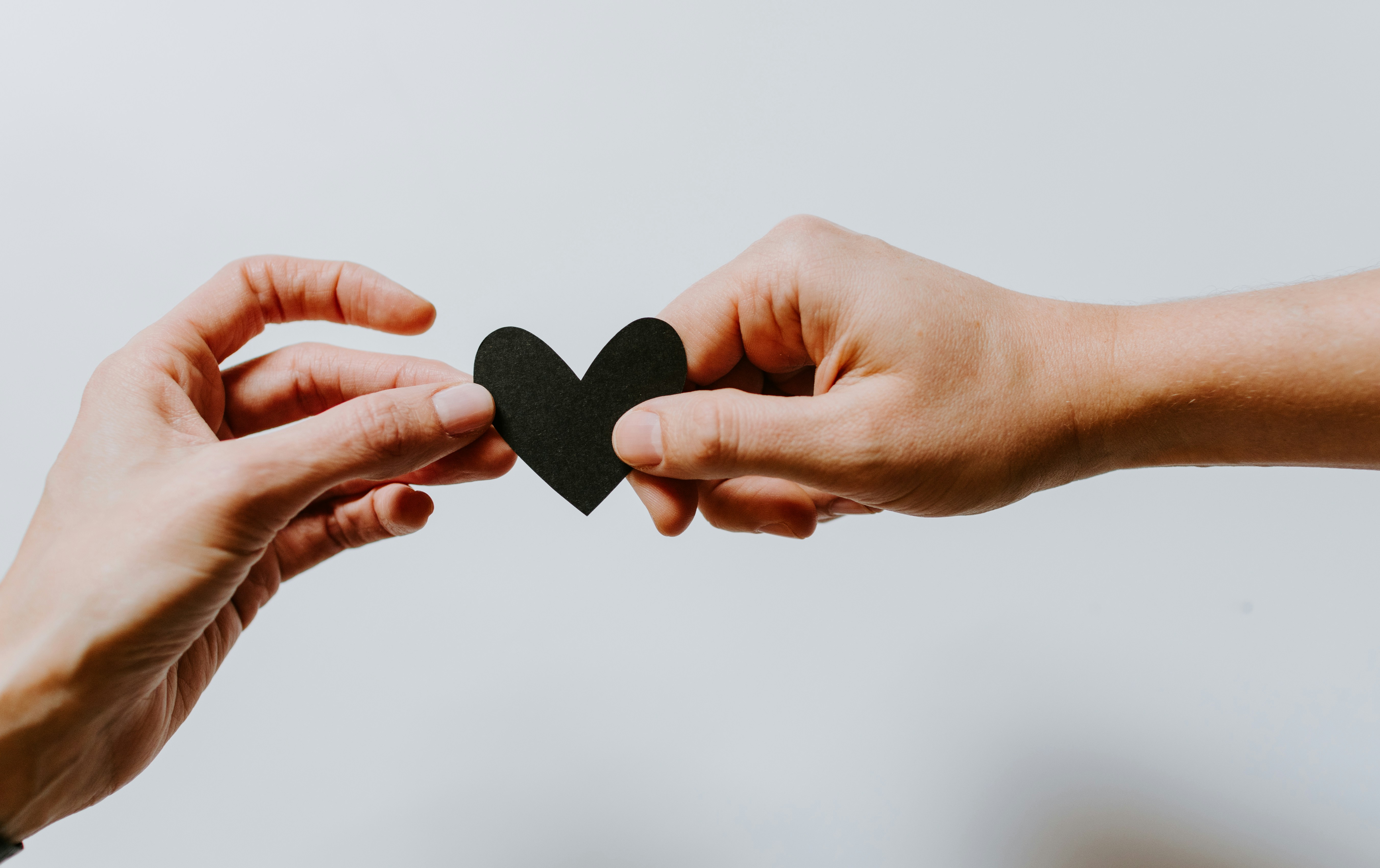 Two hands reaching toward each other to share a small black paper heart against a clean white background, symbolizing love, connection, or giving.