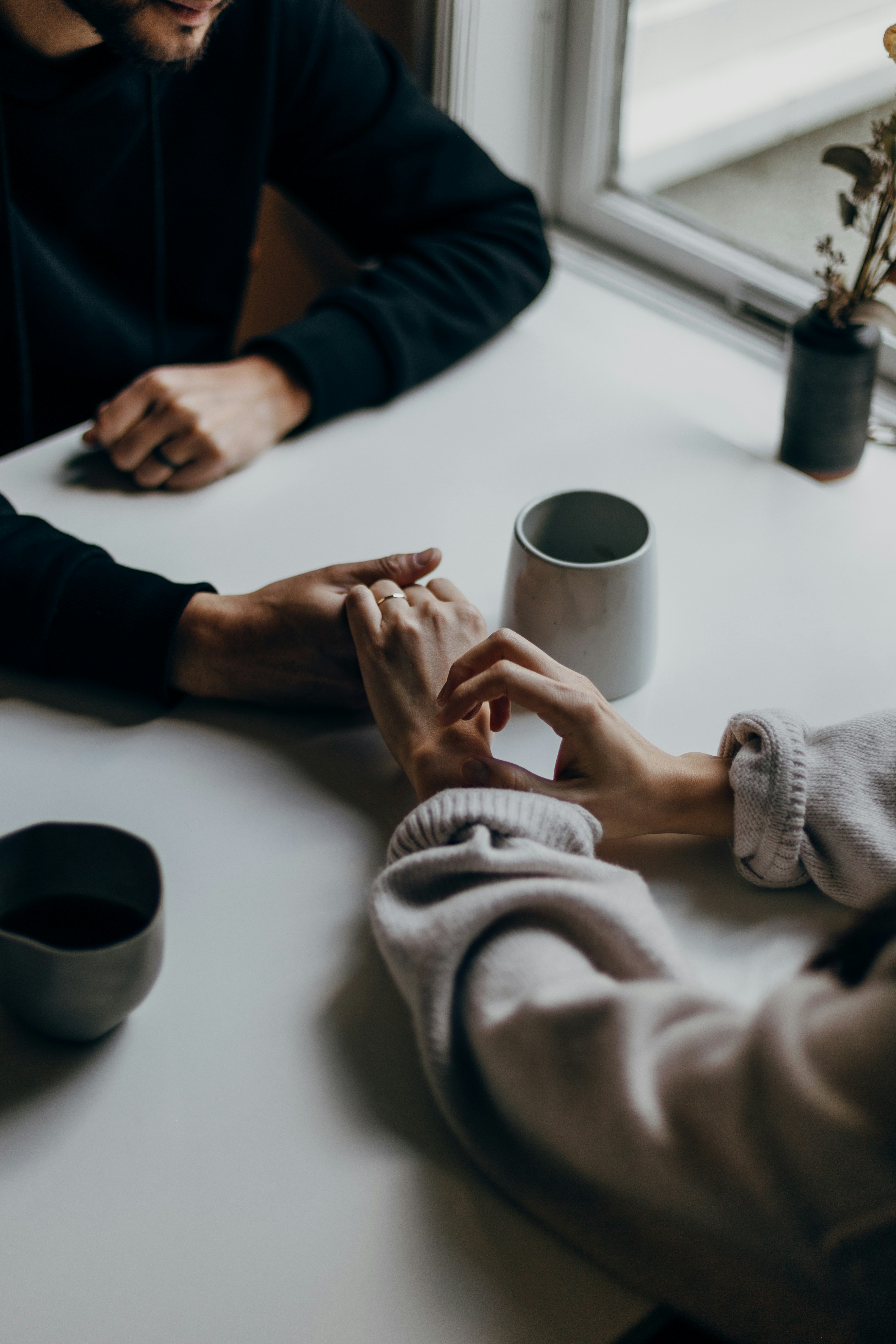 A couple holding hands across a white table during an intimate moment, with coffee mugs nearby and soft natural light from a window, one person wearing a beige knit sweater and the other in a dark top.
