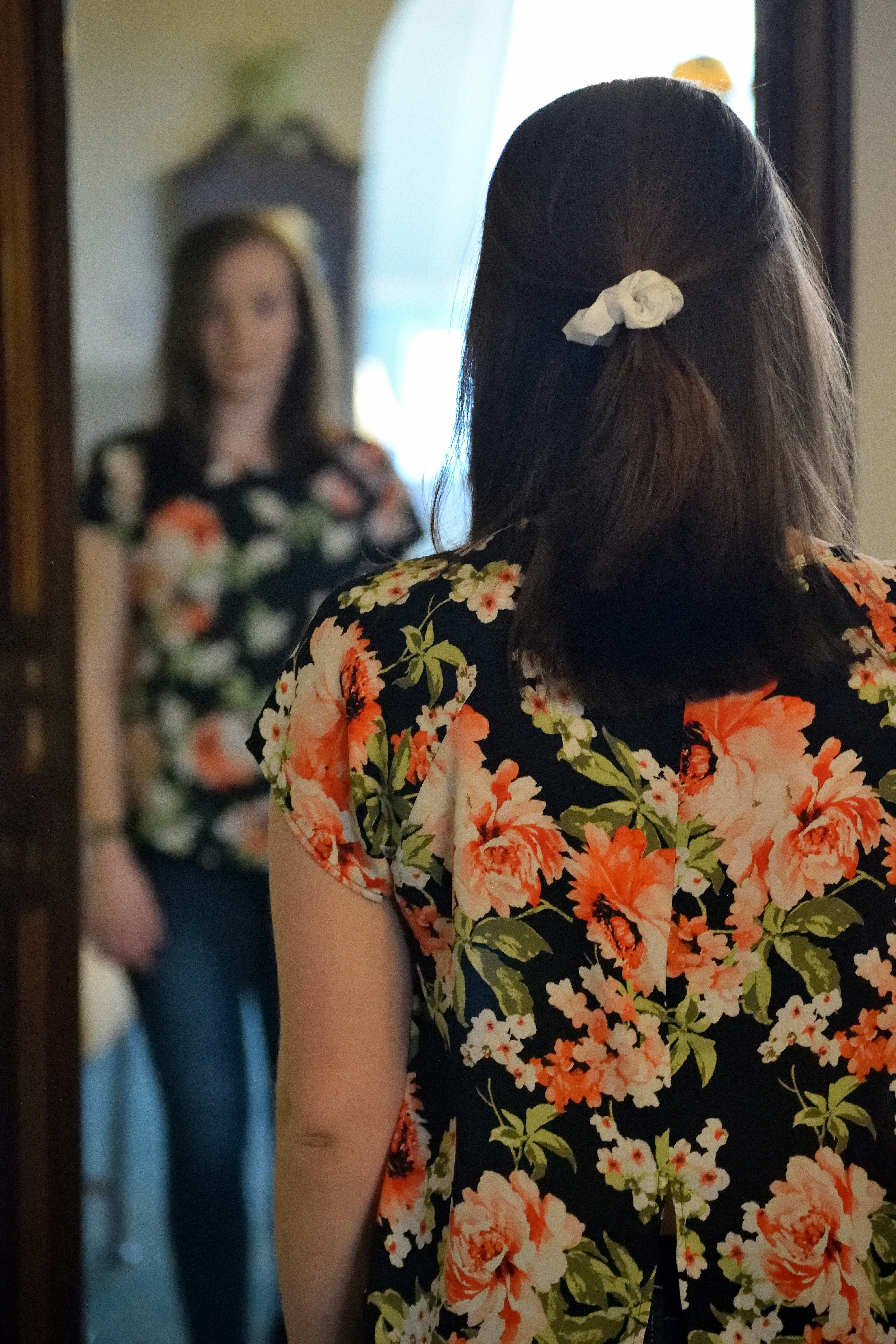 A woman with long dark hair wearing a black floral dress with orange and white flowers stands with her back to the camera, looking at her reflection in a mirror, with a small white hair accessory visible in her hair.